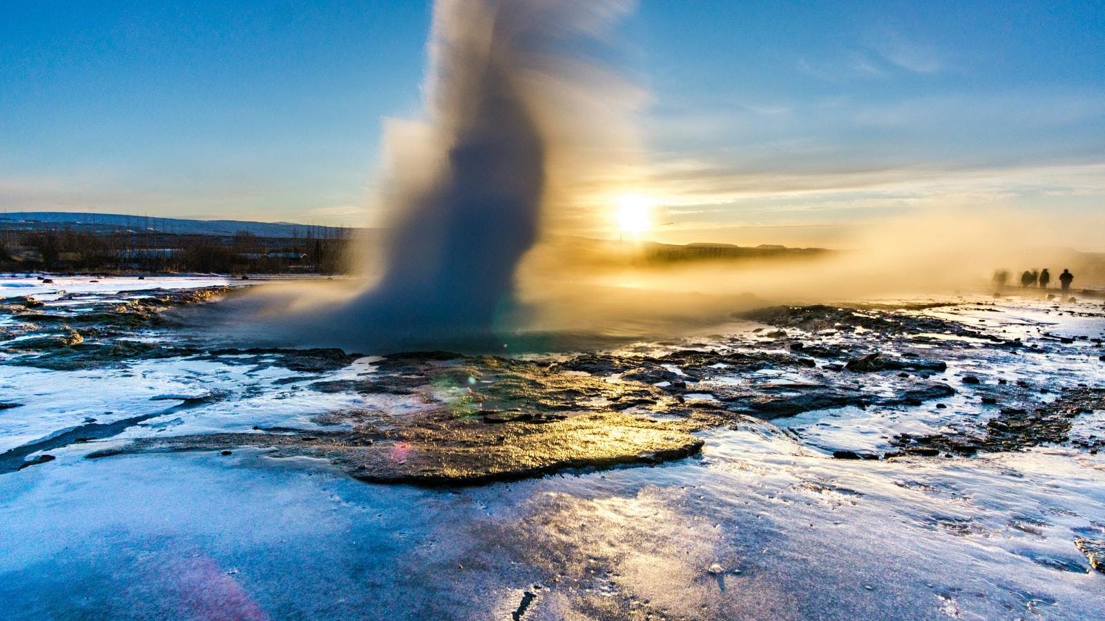 Geysir erupting on a winter day with the sun setting behind it. Tourists on a Golden Circle tour watch from a distance.