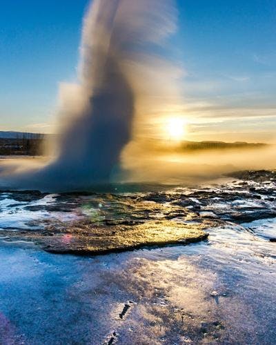 Geysir erupting on a winter day with the sun setting behind it. Tourists on a Golden Circle tour watch from a distance.